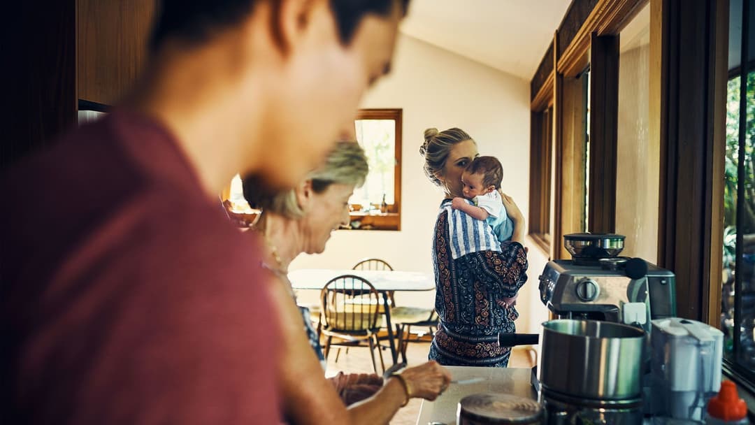 Mother holds baby in kitchen as father and grandmother make food.