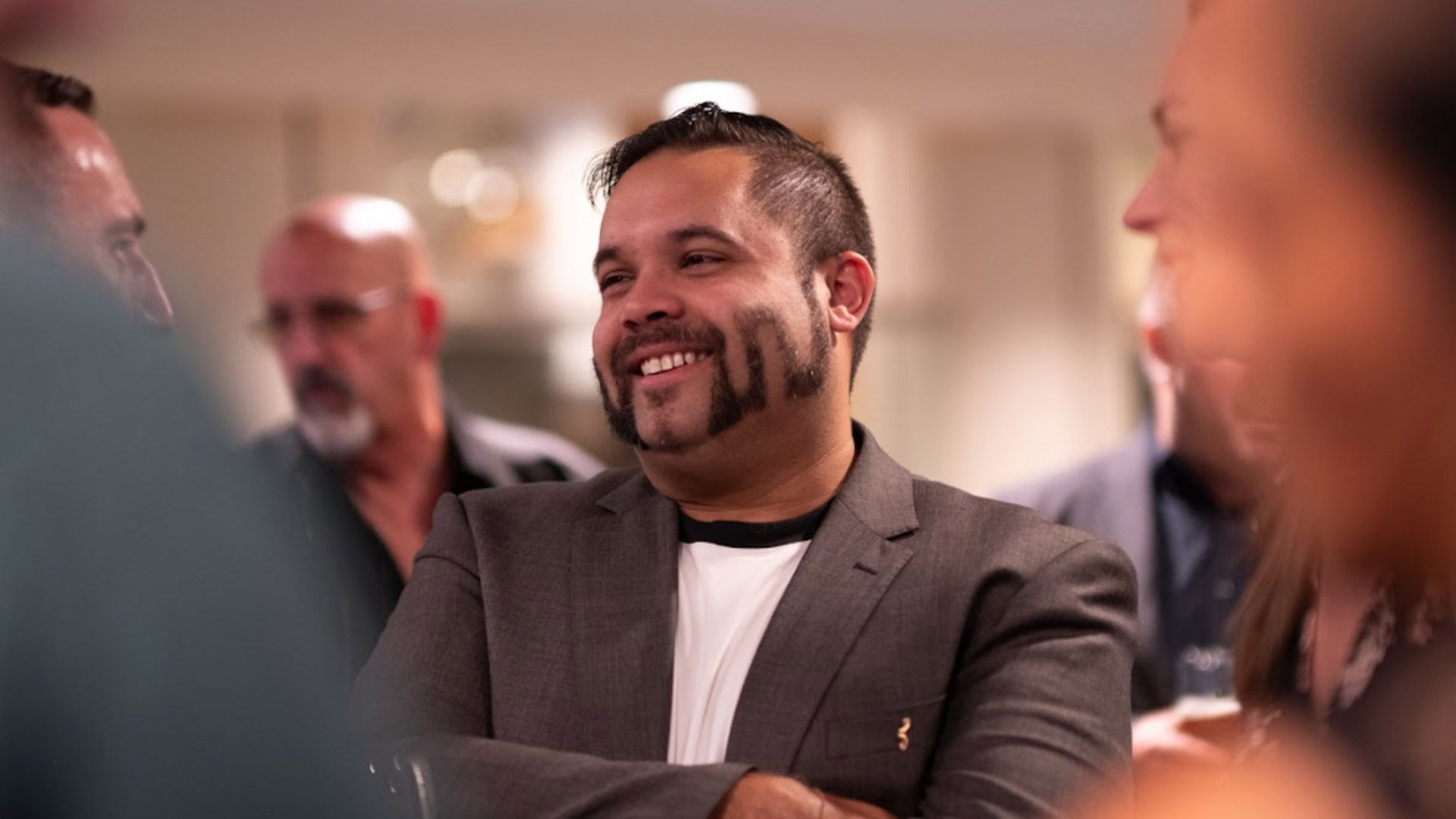 Man with unique shaved moustache design, in suit, stands with others around him at event