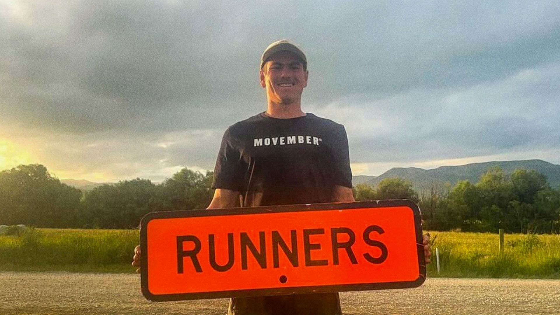 Young man stands on rural dirt road holding a street sign that says runners.