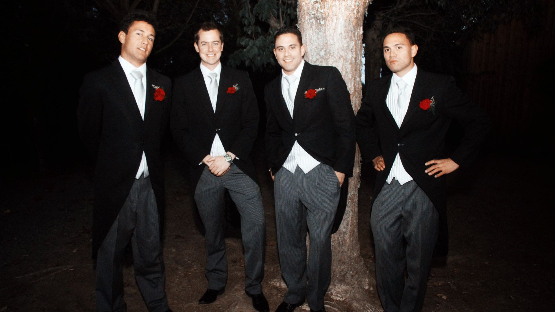 Four men in matching groomsman suits stand at night time with flash photograph