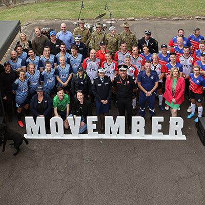 A group of uniformed first responder and army personnel, looking to camera. A Movember sign is in the foreground.