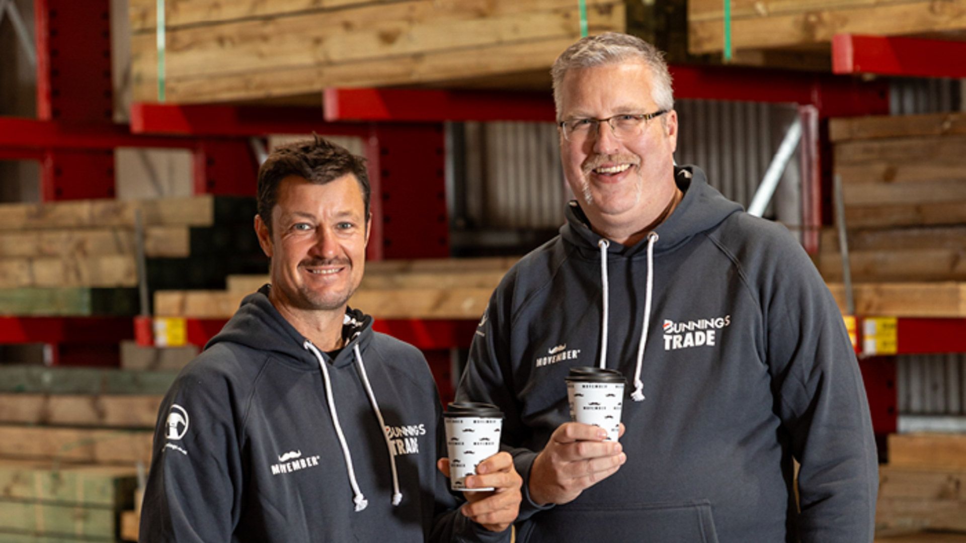 Two men in green hoddies and coffee cups with timber in background of trade store
