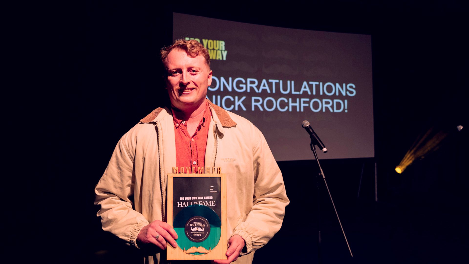 Man holding Movember award on stage, mic to the left, background his name appears on screen