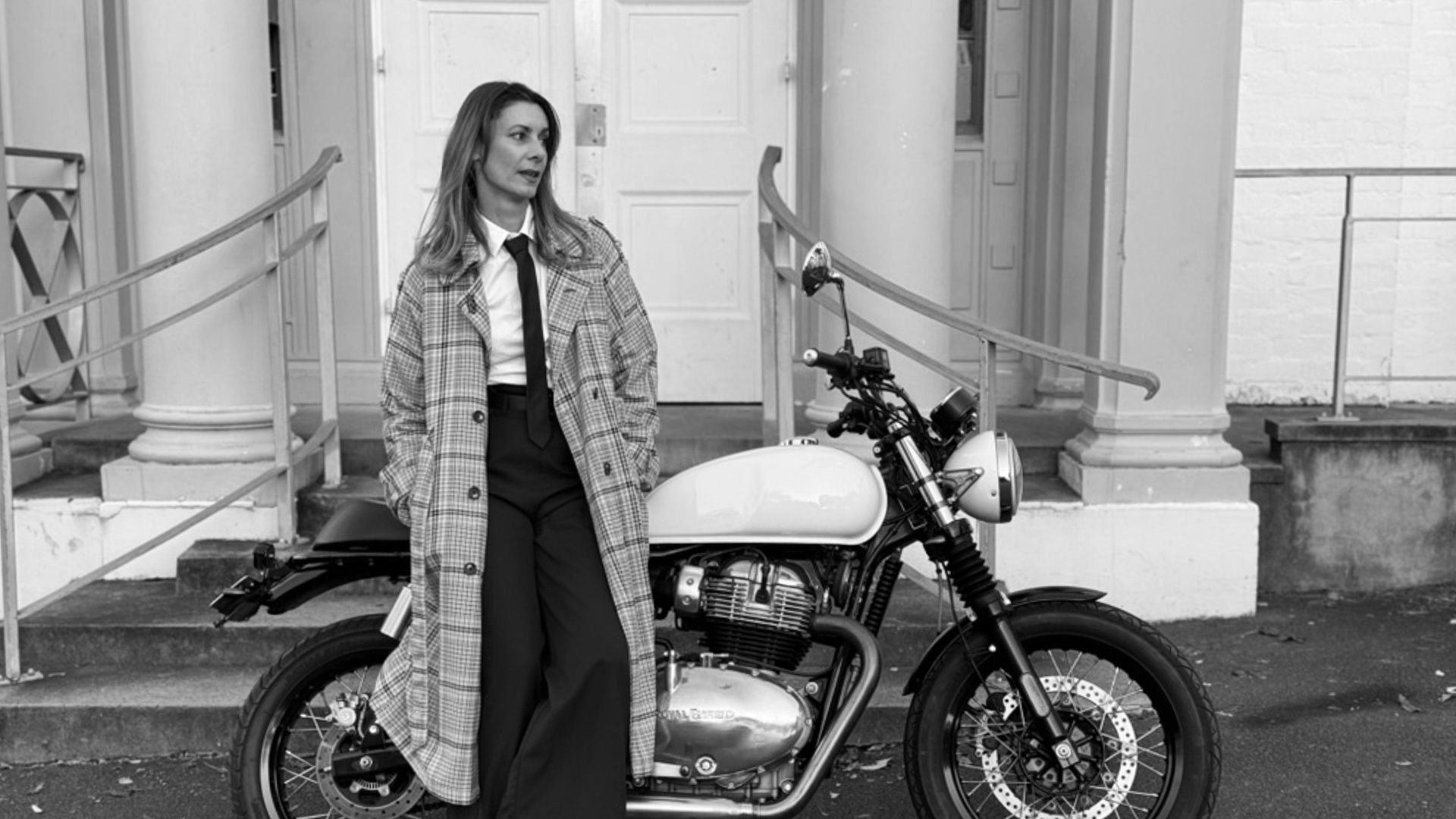 Woman in coat stands next to Royal Enfield vintage motorbike, parked against white wall. Black and white photo