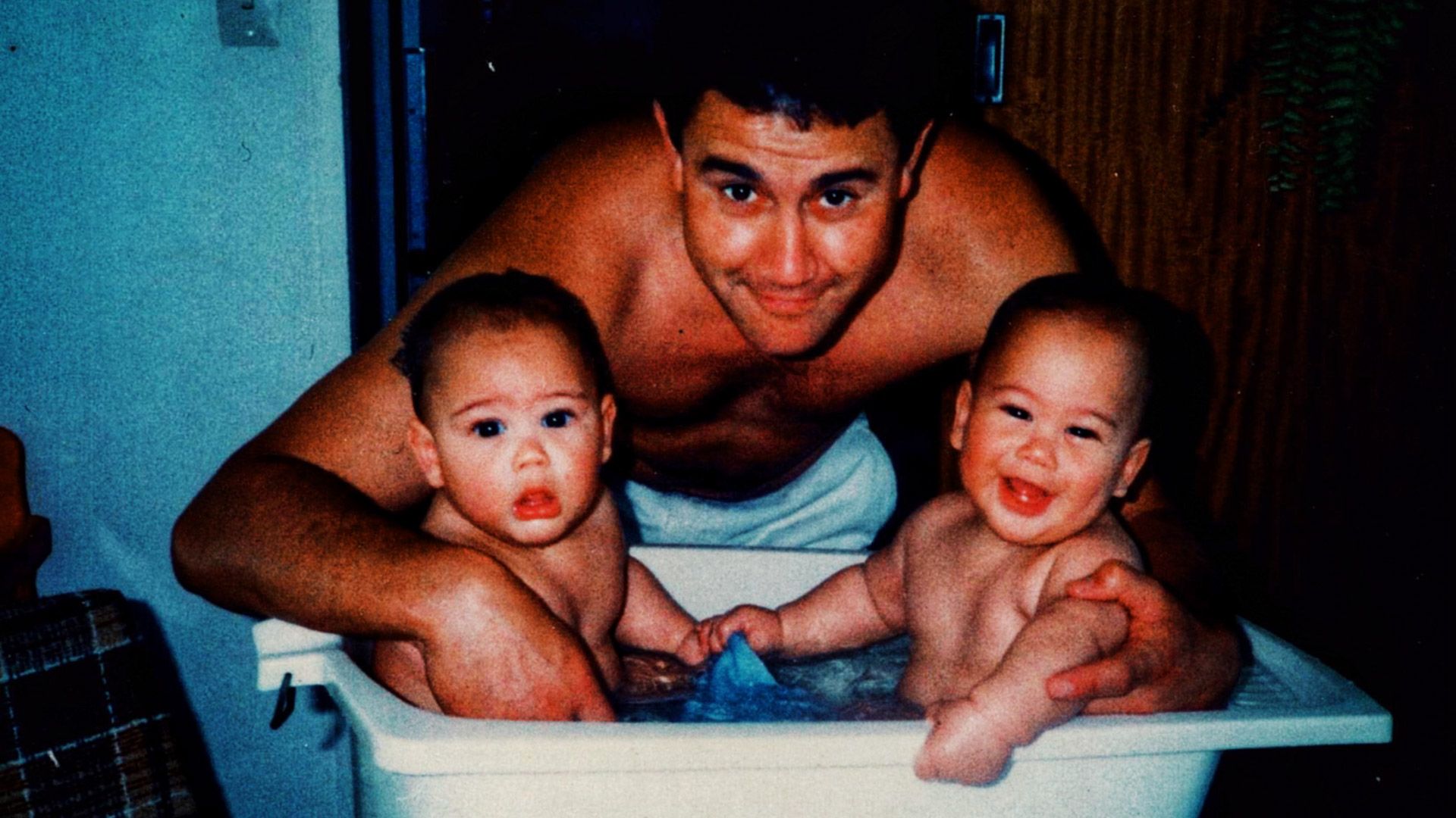 Two babies in small bathtub with adult man standing above them smiling to camera