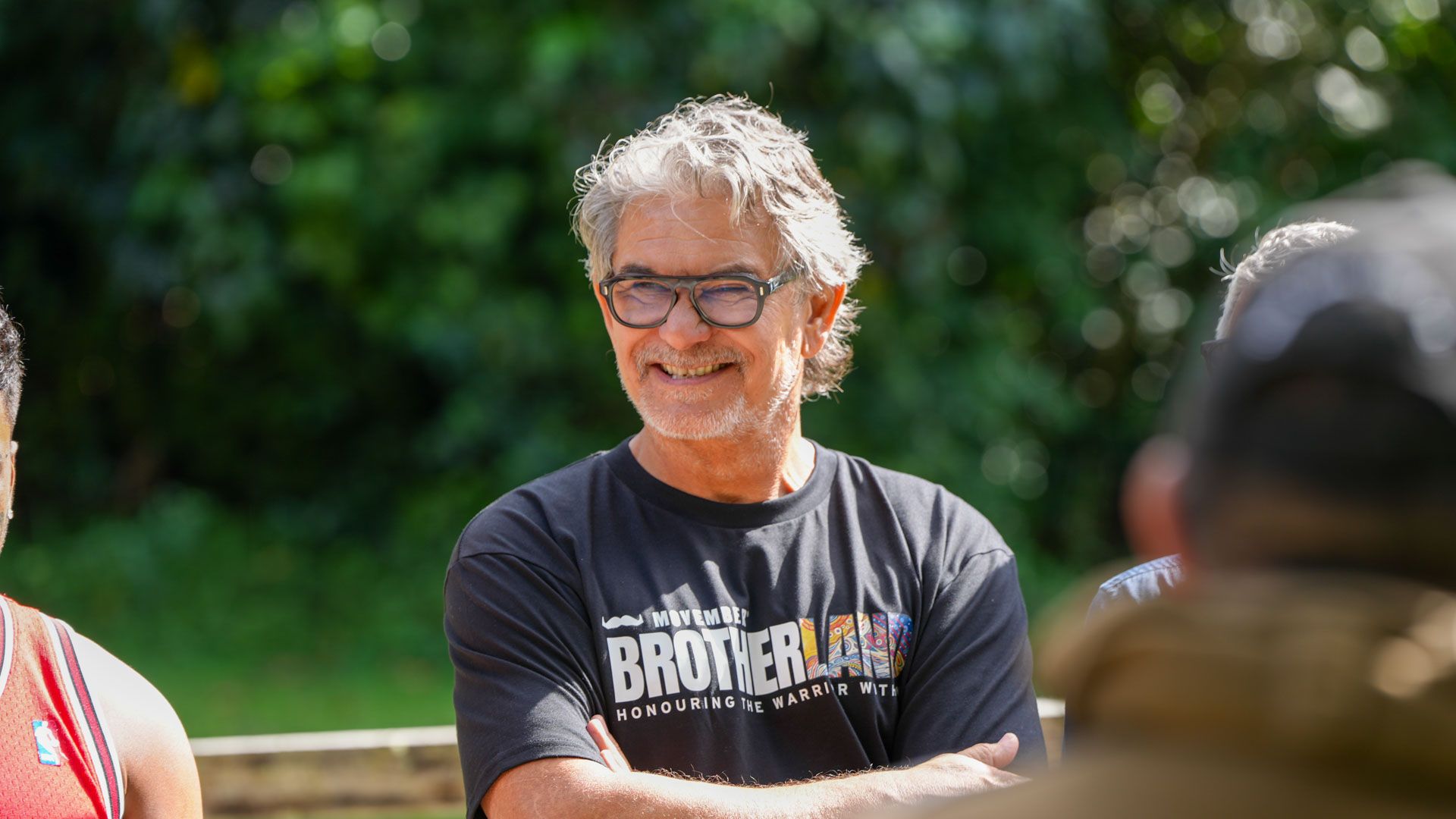 Man iwth glasses and tshirt saying 'brotherland' folds his arms and smiles to someone off camera