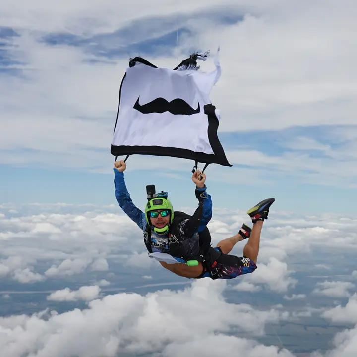 Skydiver looking to camera while descending from the sky and holding Movember flag