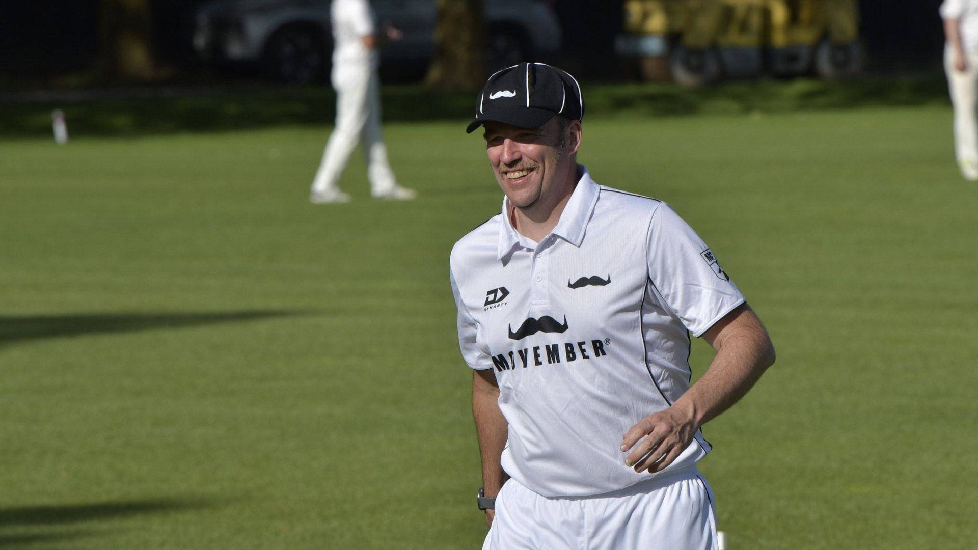 Man with moustache wearing cricket whites and black hat running on cricket field