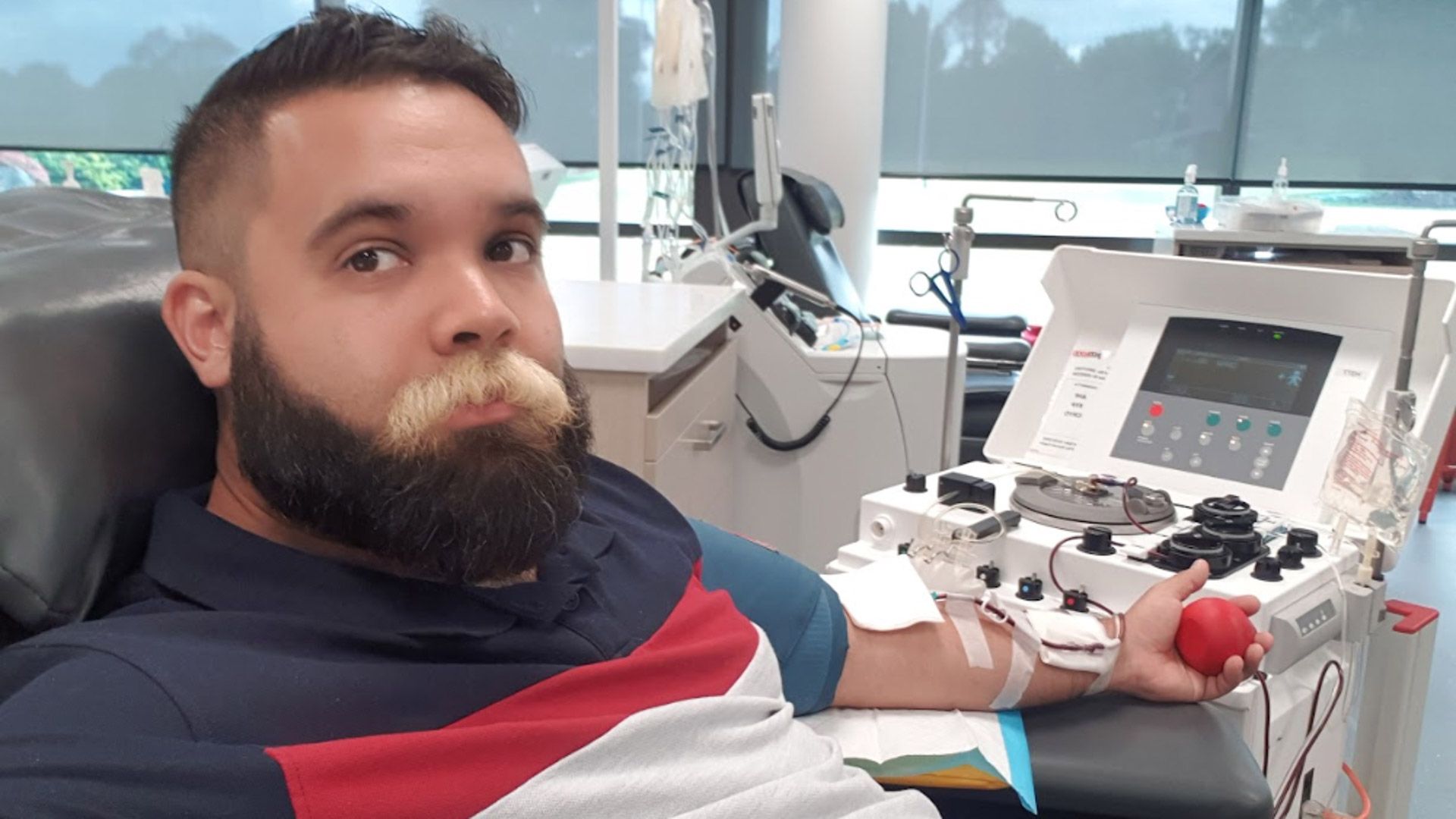 Man with blonde moustache and brown beard sits on blood donation chair taking selfie holding red squish ball