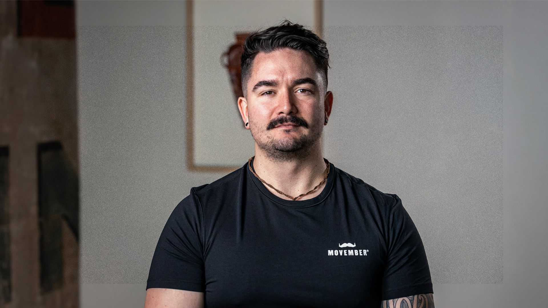 Man in movember t-shirt stares at camera stoically, within a room with grey walls