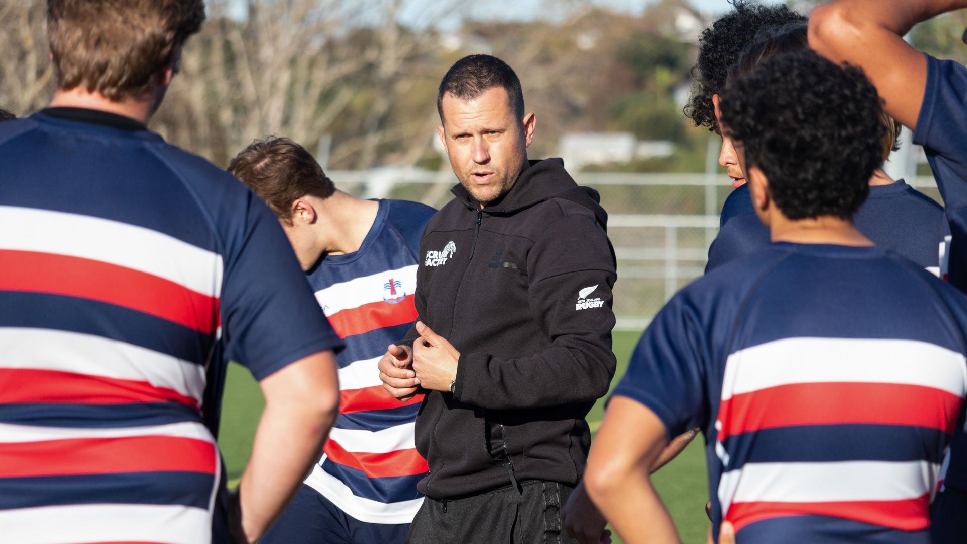 Rugby coach stands on field surrounded by teenage players in striped uniforms