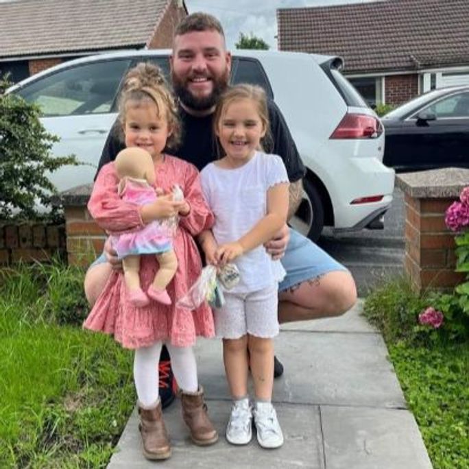 Photo of man posing in a driveway, beaming while embracing his his two daughters.