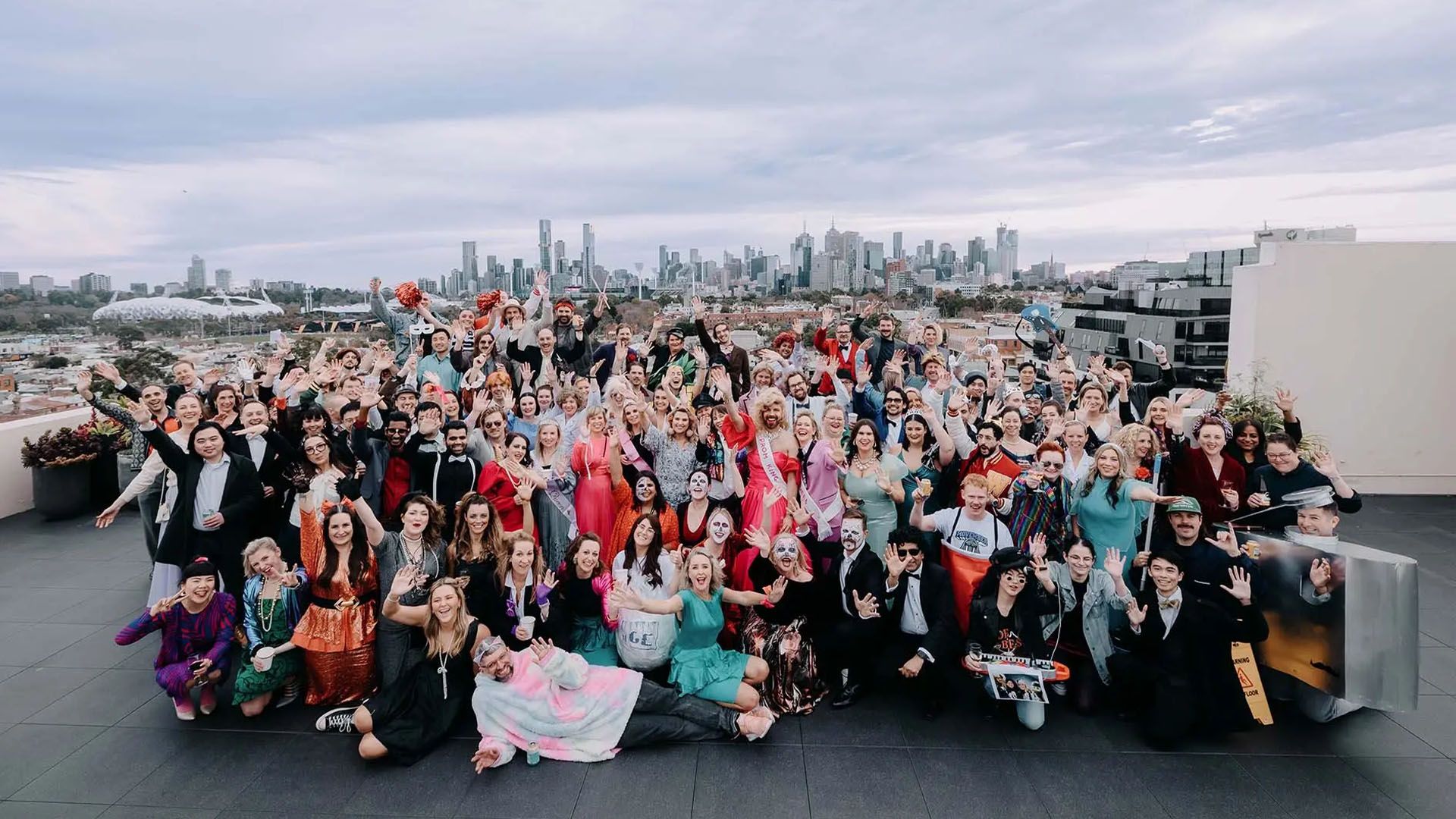 Photo of a large crowd of people on a rooftop, dressed in themed costume. The Melbourne Cricket Ground and Melbourne skyline is visible behind them.