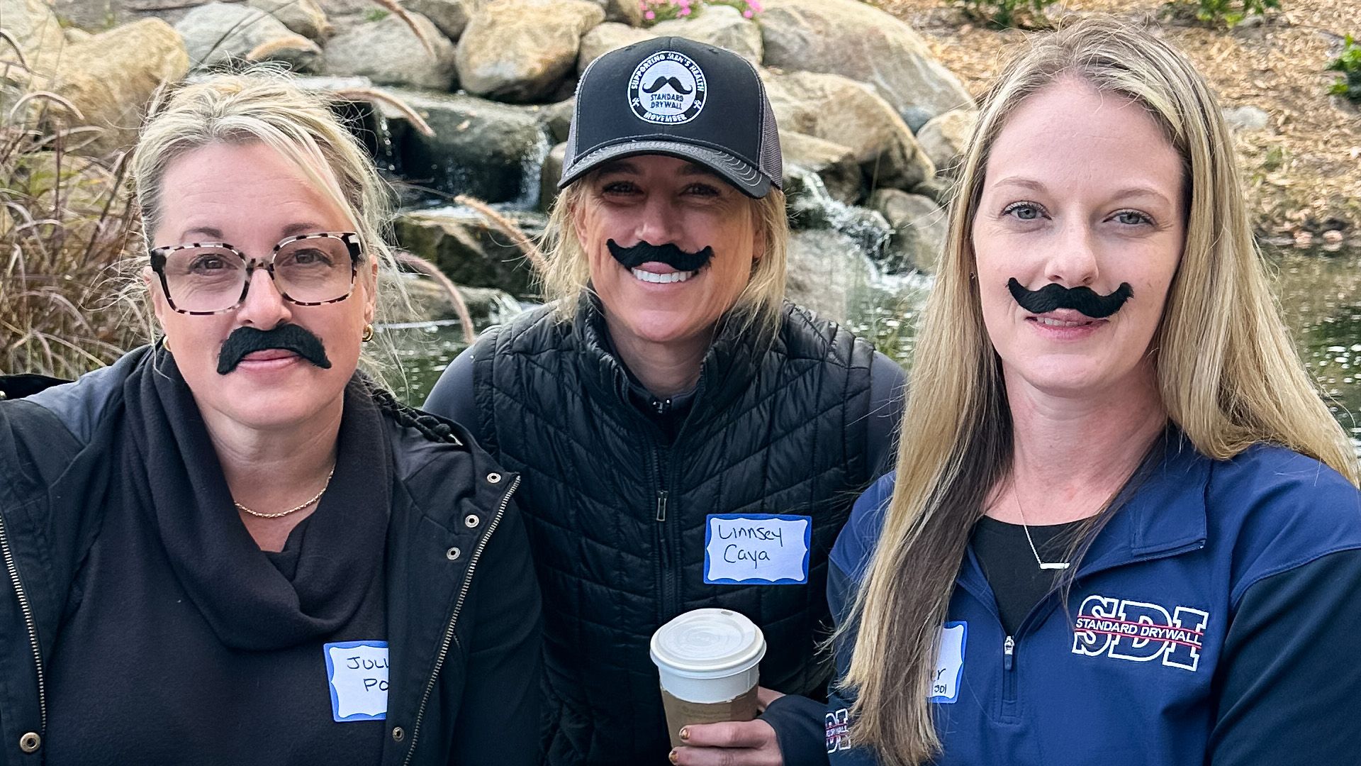 Three women smile together, each is wearing a funny stick-on moustache