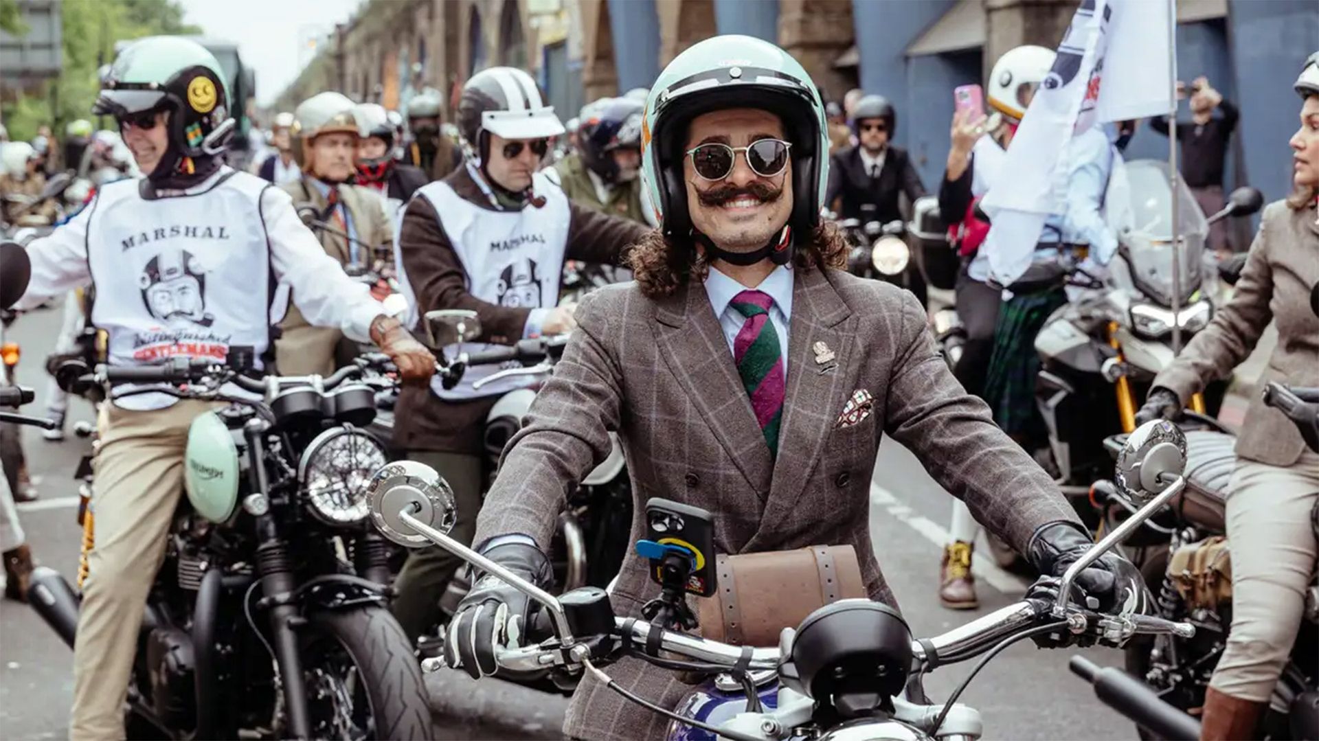Photo of a dapper man with a magnificent moustache, riding a classic motorcycle.