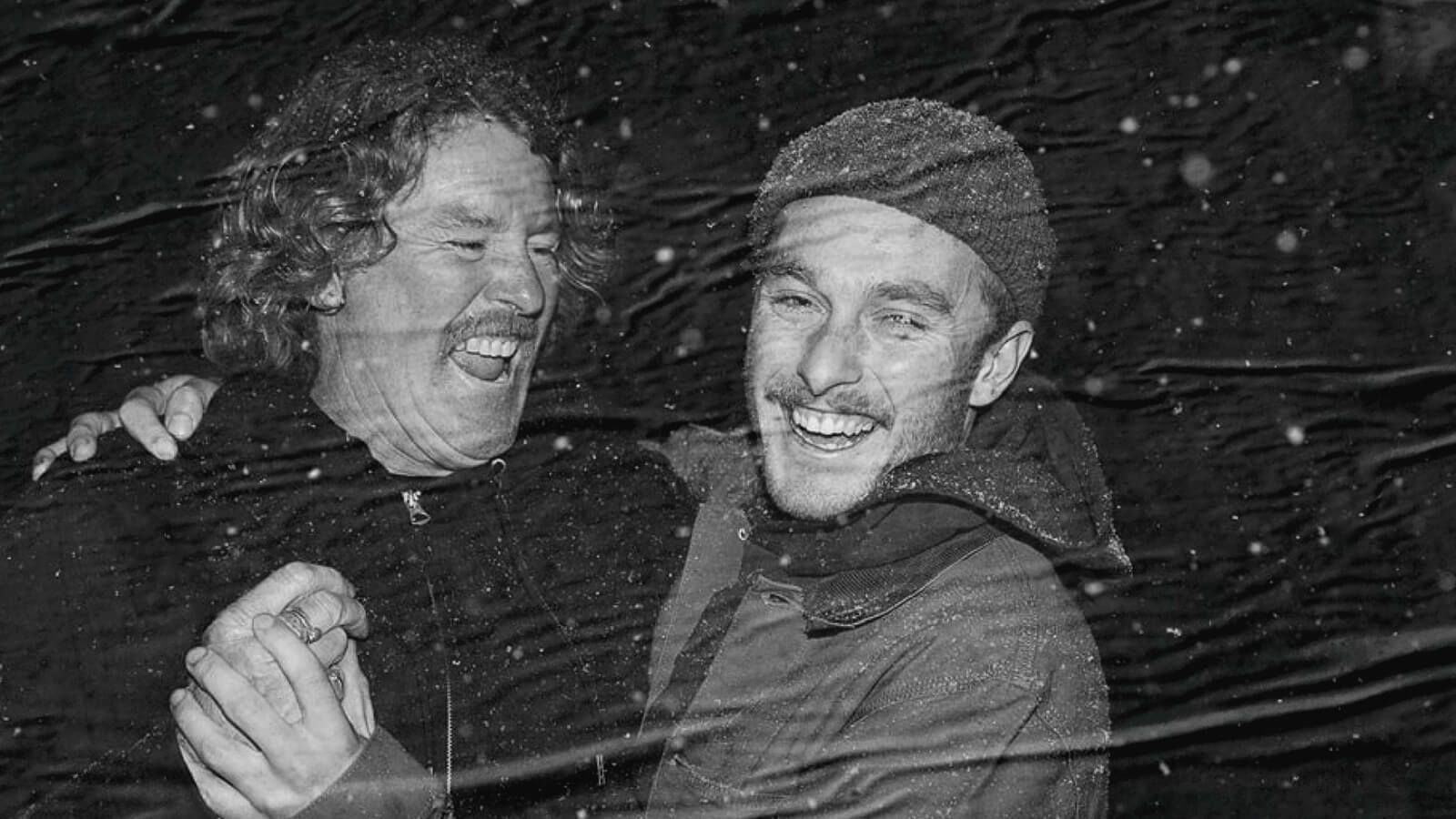 Two men, smiling wildly to camera, semi-embracing under a light snowfall.