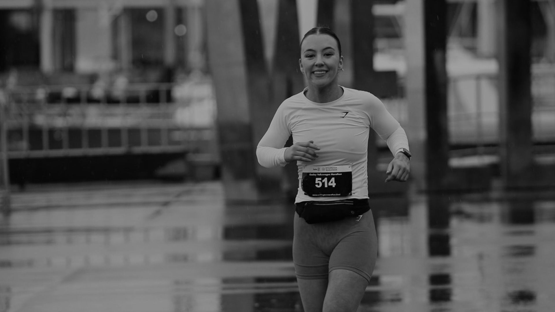 Black and white, woman running on waterfront with run event number on top, looking and smiling at camera