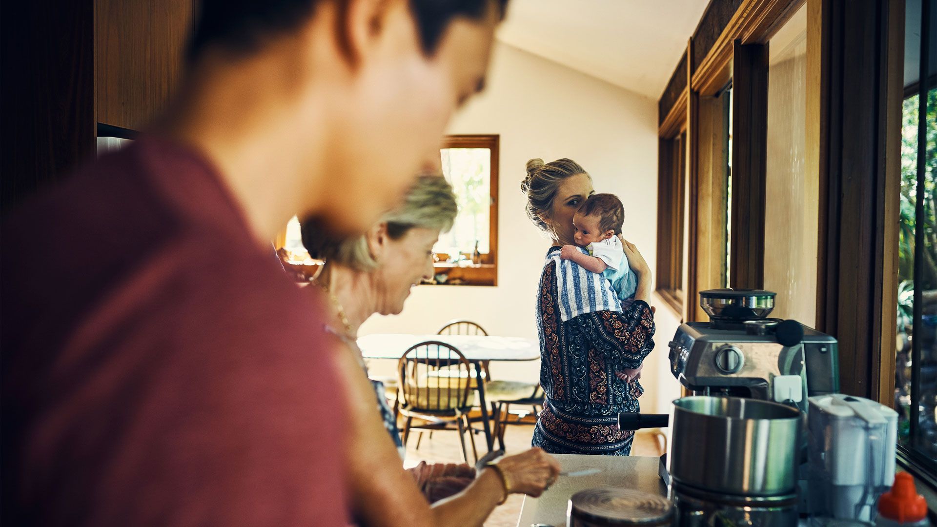 A photo of a mother holding a baby in a kitchen as a father and grandmother make food.